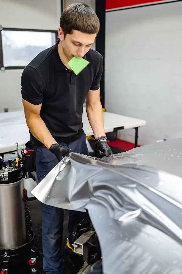 Gros plan sur le visage d'un spécialiste de la pose de film chez PISTA, tenant une raclette de finition verte entre ses lèvres tout en inspectant minutieusement l'application d'un film PPF gris mat.