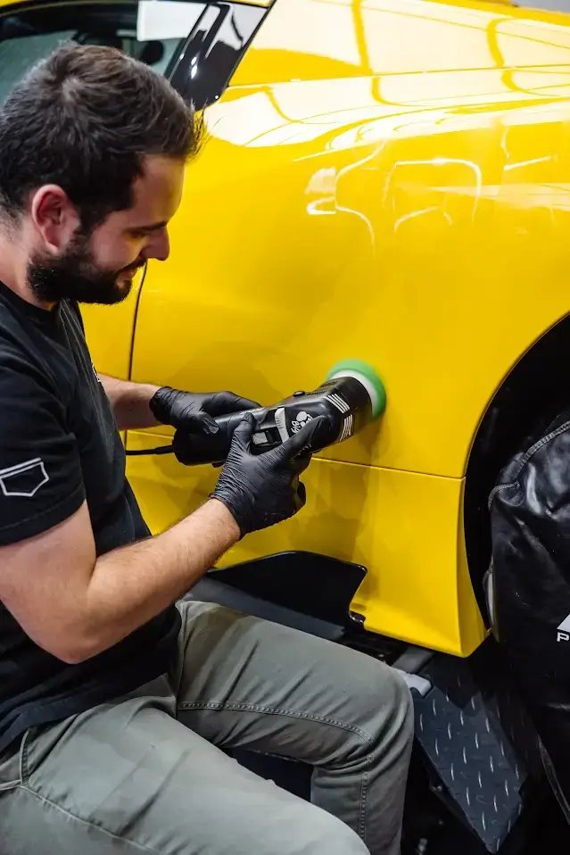 Un technicien PISTA spécialisé en correction de peinture, portant des gants noirs, effectuant un polissage de précision sur le bas de caisse latéral d'une voiture de sport jaune.