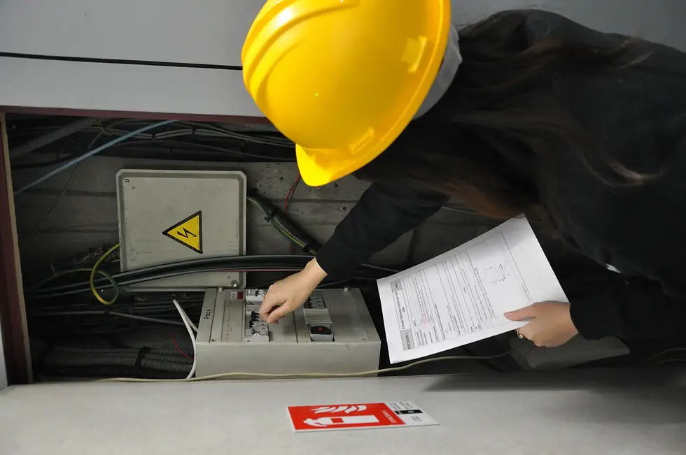 Technicienne en casque jaune inspectant un coffret électrique ouvert avec un plan à la main, en milieu industriel.