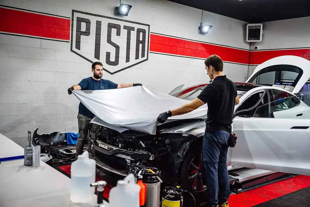 Deux techniciens de l'atelier PISTA, portant des gants de protection noirs, appliquent avec précision un large film de protection carrosserie (PPF) de couleur gris mat sur le capot d'une voiture de luxe blanche partiellement démontée.