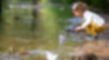 Young toddler girl looking at a paper boat on a creek.