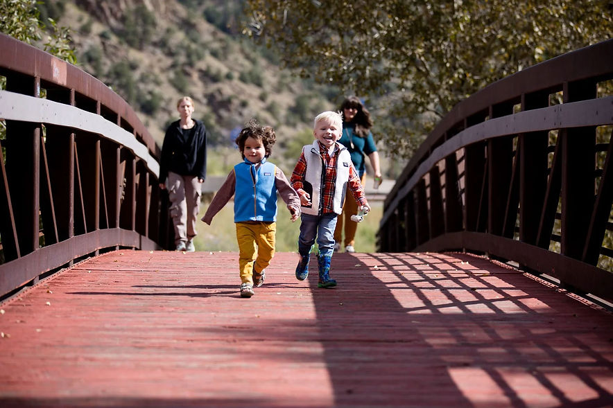 Two kids running over a bridge with two female Mountain Trails daycare providers walking behind them in Clear Creek county Colorado.