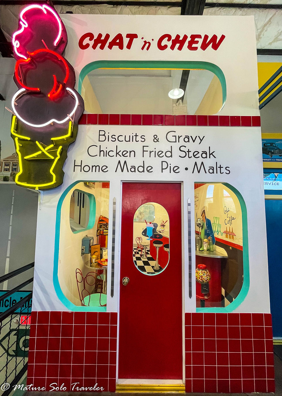 a replica of a 1950s diner. The sign says it is called the Chat 'n' chew. It advertisies biscuits and gravy, chicken friend steakk, home made pie and malts. a neon ice cream cone lights up on the side and red checkered tiles are on the bottom third of the wall. through the centre red door black and white floor tiles can be seen below a lunch counter in the diner.