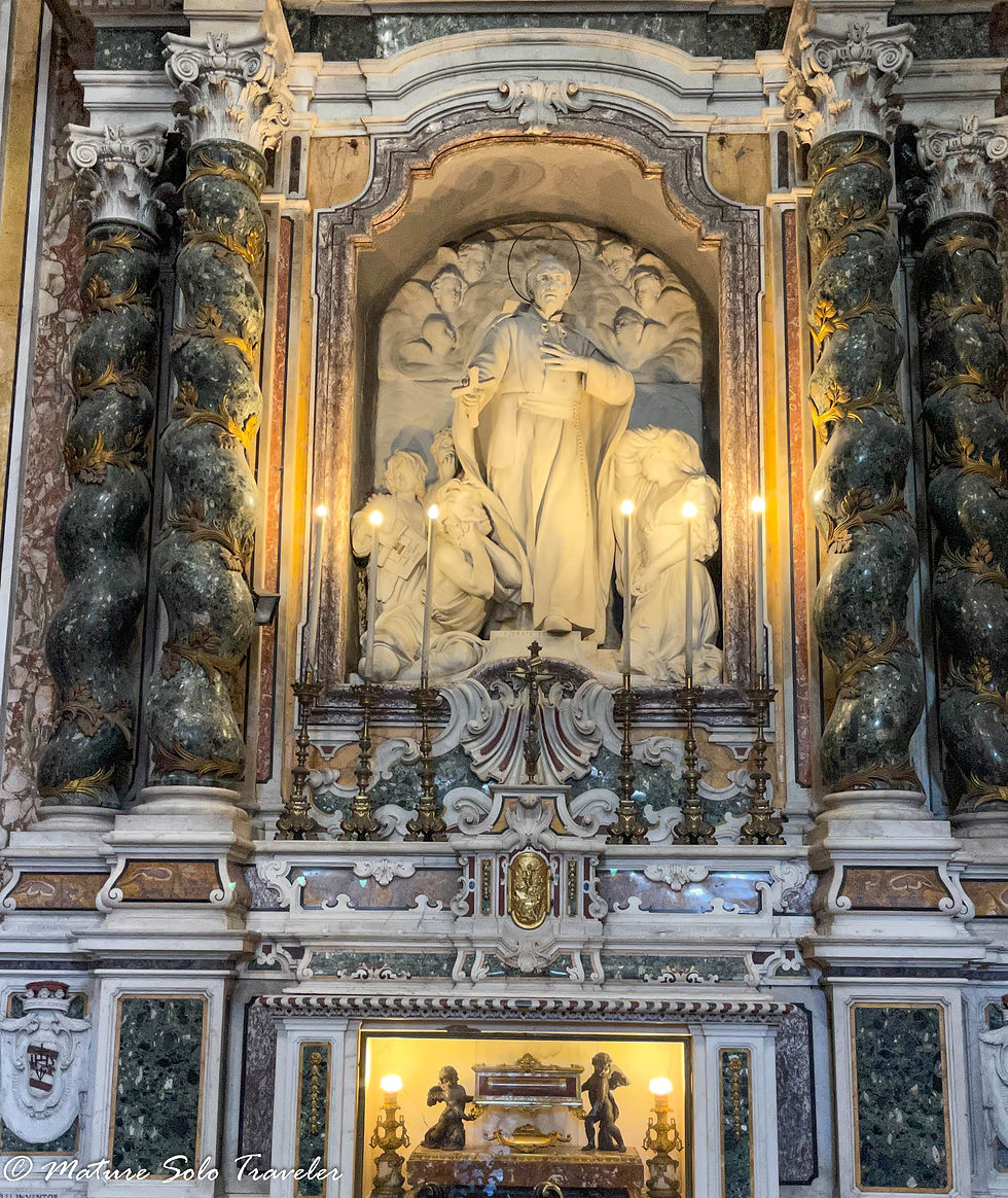 Upon an altar is a life sized statue of a priest with a halo around his head. A despairing woman reaches for him on his right and two young men beg for a favour on his right. Above his head in the clouds are just barely visible little cupids. The whole setting is above an alter with twisted black marble columns on each side and an effigy of a coffin behind glass below it.