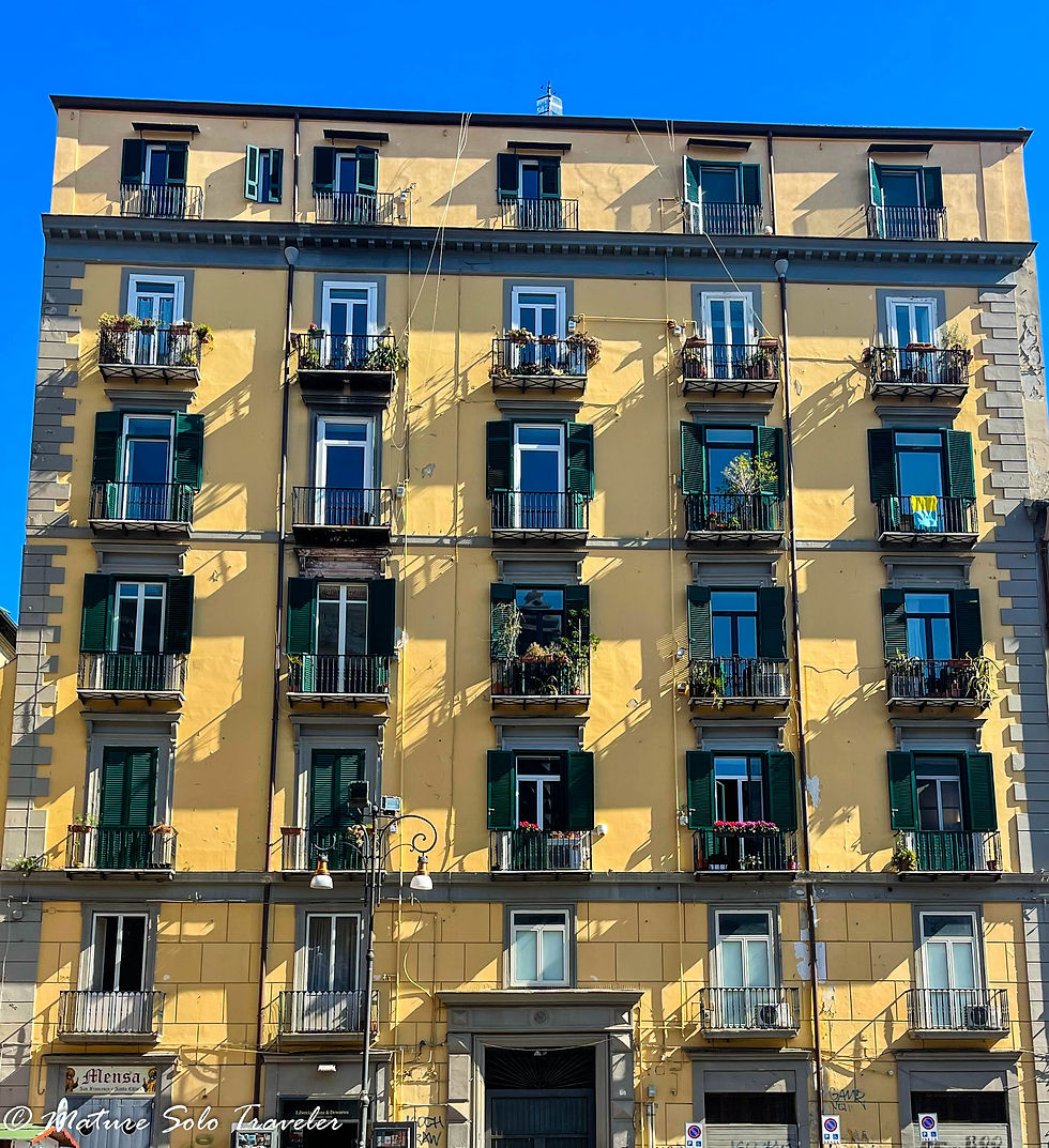 a seven story apartment building. yellow walls with five tall windows across each level. each window has a little metal balcony. most of the balconies have pot plants on them.