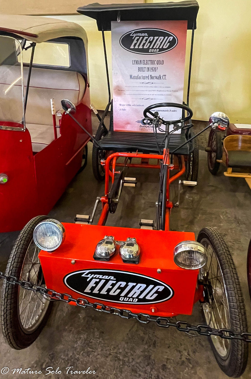 this Lyman electric quad car was possibly built in 1970, manufactured in Norwalk CT. It looks like a child's cart, with bike pedals for both the driver and passenger. they both sit on an open seat, with a cover above their heads. There is no body on this electric vehicle