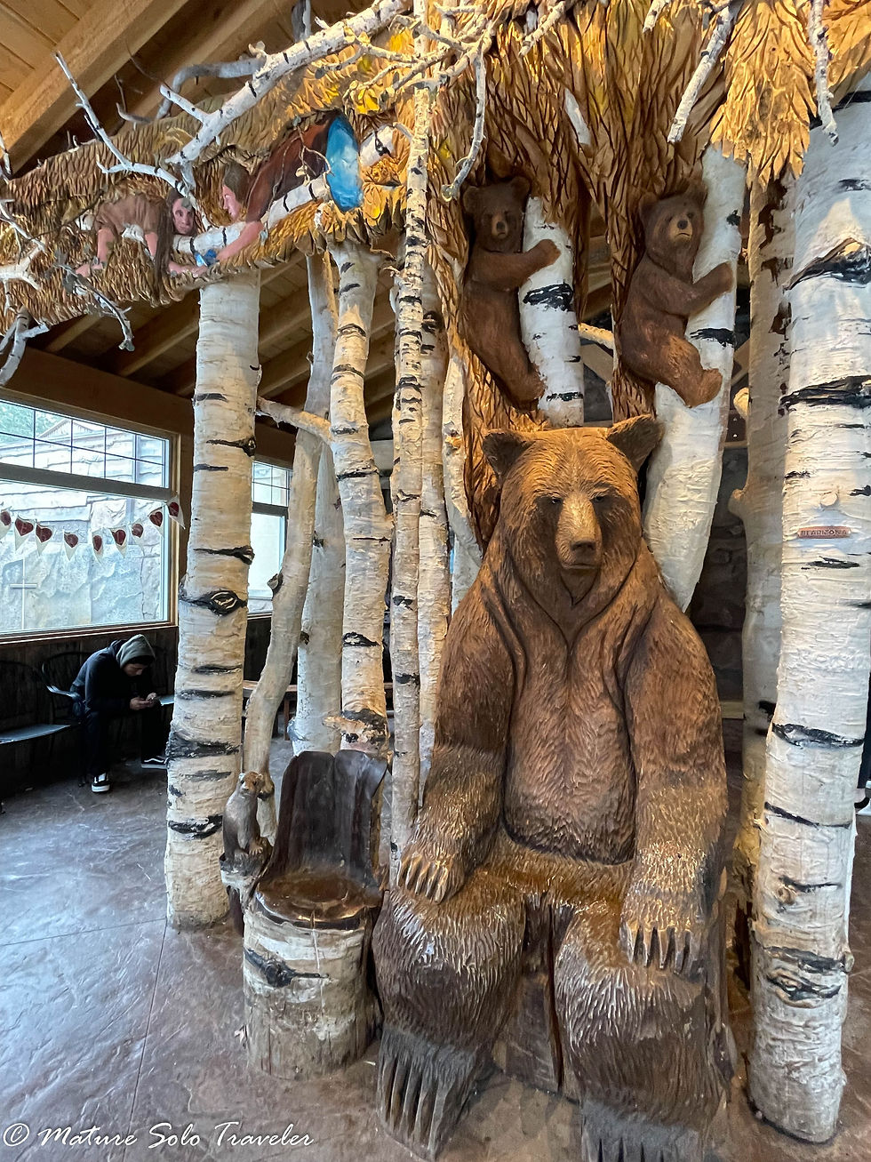 the LaBenne family have sculptured a grizzly bear sitting down and visitors can take a selfie while sitting on his knee. two grizzly bear cubs climb the trunk above their mumma