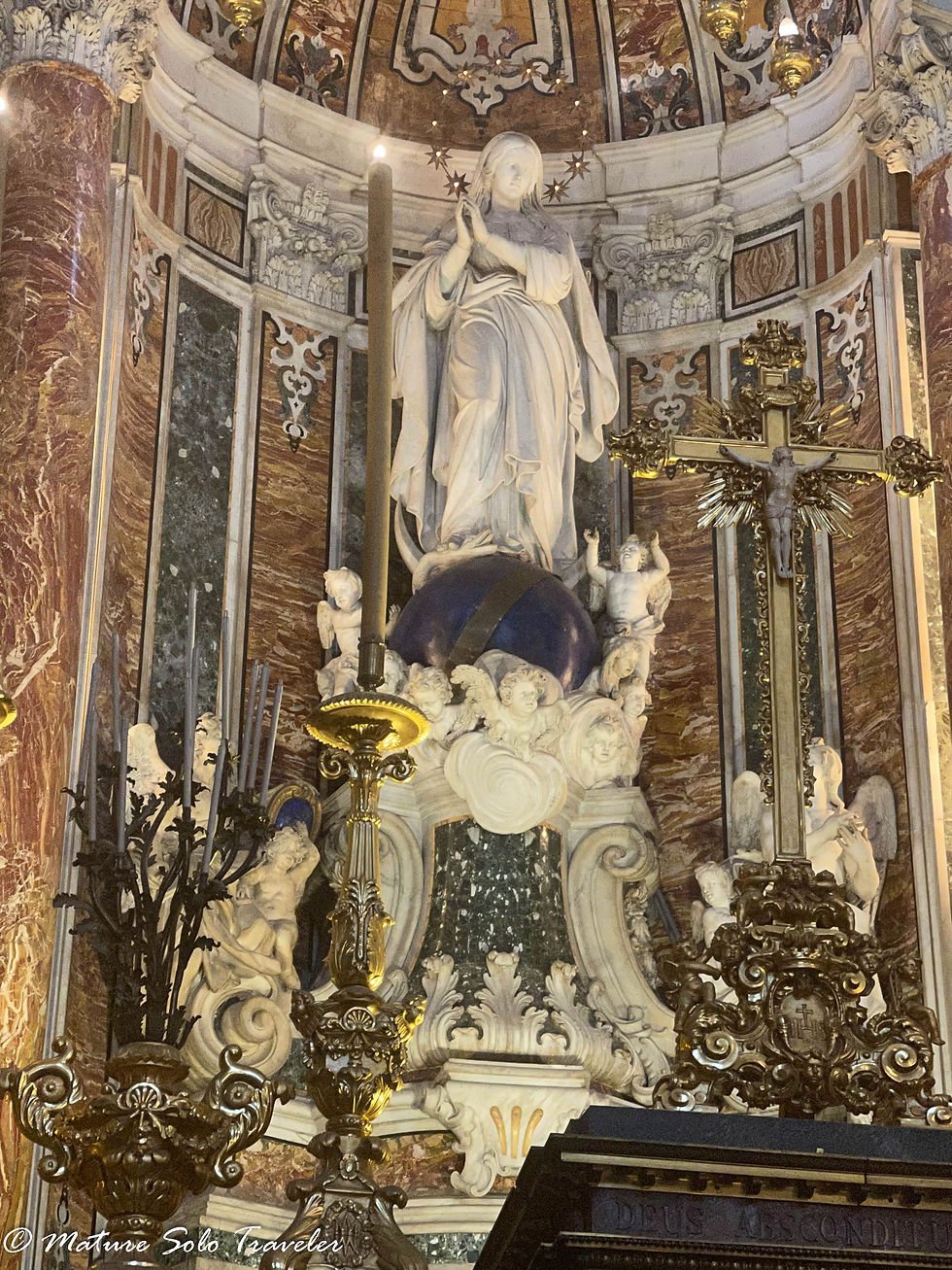 the Virgin Mary stands atop a blue globe of the world, a circle of stars surrounding her head like a halo. This marbe statue is surrounded by cherub angels looking up at her. The marble decorated walls form an alcove around her body, and highly decorated plaster plinths in white contrast with the coloured marble.