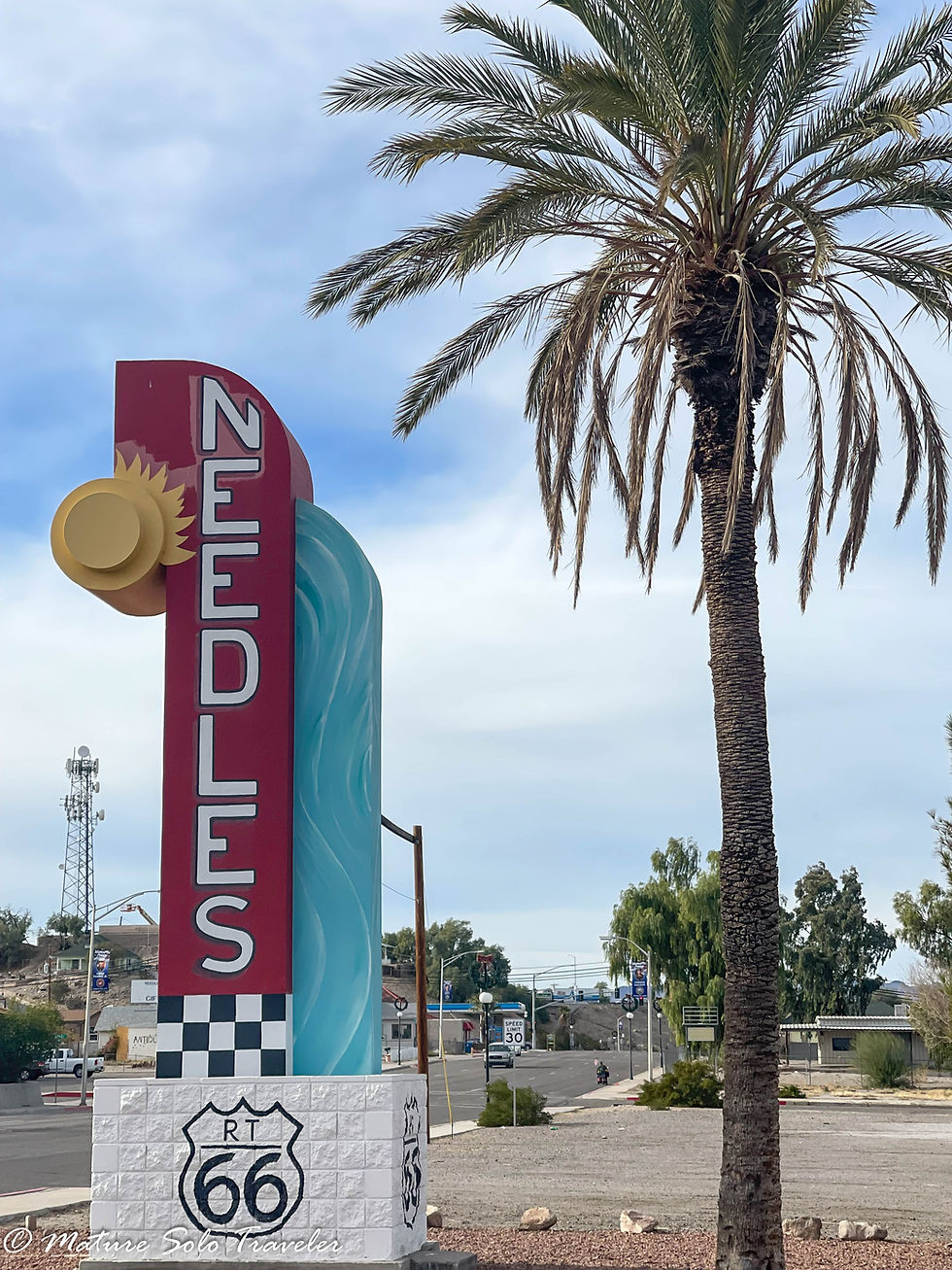 a large sign stating Needles rises above a white painted brick base with the route 66 shield painted on all 4 sides. The needles sign is red with white writing, andhas a yellow sun near the top. To the right of the sign is a single palm tree, taller than the sign