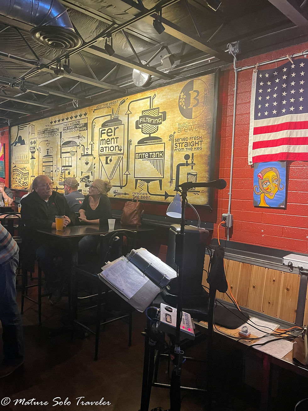 At black bridge brewery, the wall are decorated showing the process of how they make their beer. A music stand and microphone are ready for the musician to entertain the crowd. People sit at high stools enjoying their evening out.