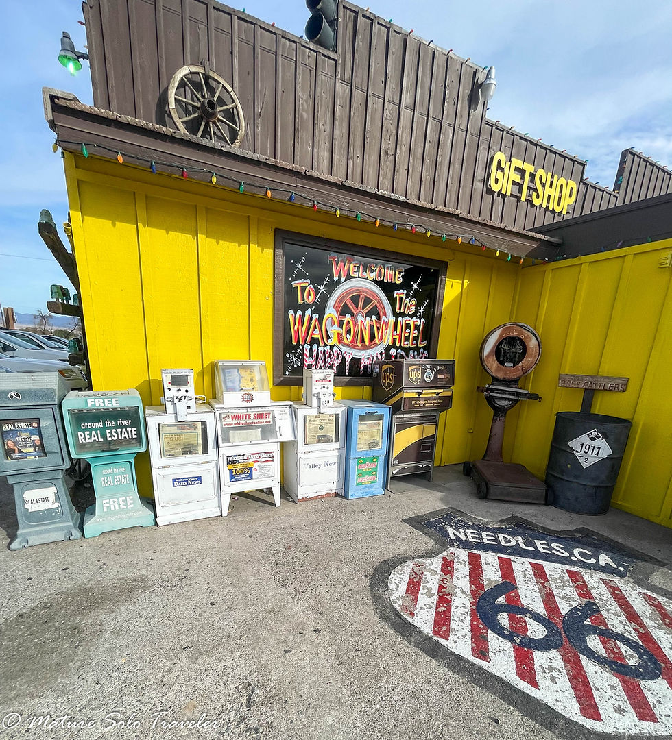 an external shot of the Wagon Wheel restaurant. The bottom floor of the building is painted a bright yellow.. seven newspaper despensers stand against the wall. a route 66 shield is painted on the concrete outside. It has needles written at the top of the shield. and the 66 is painted over a red and white striped base.