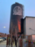 A multi story black painted beer brewer has the rickety cricket brewery logo in white on it. A ladder leads up to the top. Along the fence down the bottom are strung colorful christmas tree lights.