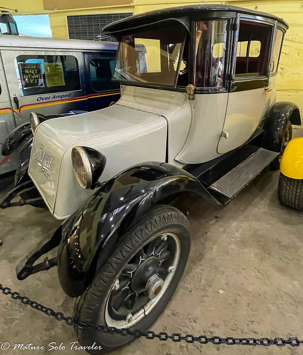 this Detroit electric car looks similar to an early model ford. its front windscreen opens from the bottom and pushes out. its paintwork is grey, and it has black hubs over the wheels. there is a running board on either side of the car.