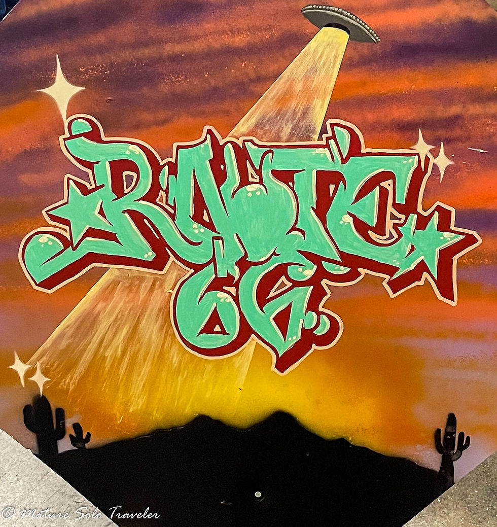 a graffitied decommissioned street sign features a UFA beaming down on a desert landscape. the words Route 66 boldly emblazzed in green and red paint across the middle.