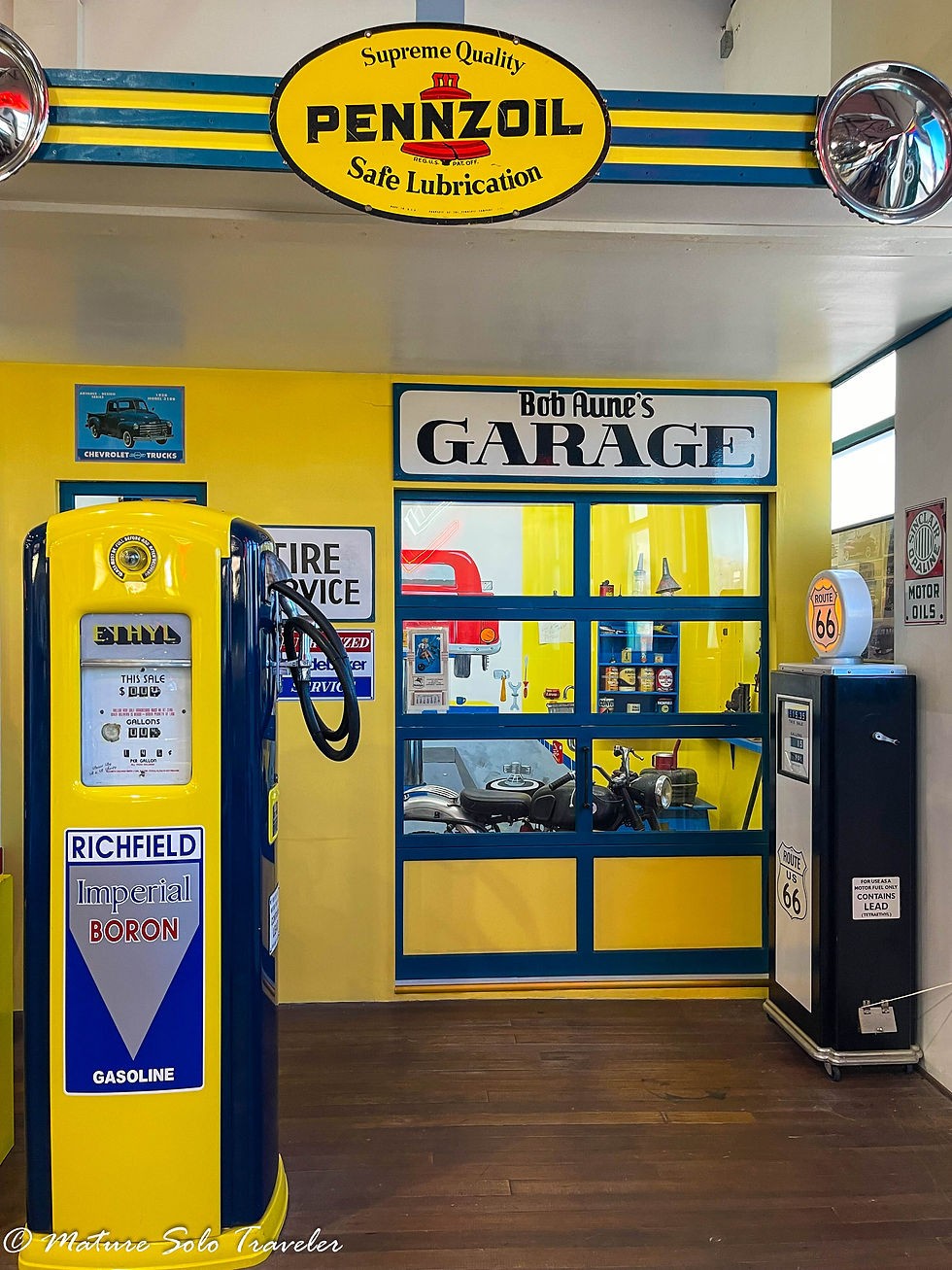 a mock up of a garage from the 1950s era. Pennzoil safe lubrication sign hangs from the roof. Two gas pumps from the day stand in the forefront. The walls of the garage are painted yellow and a vehicle can be seen through the glass door of the garage.