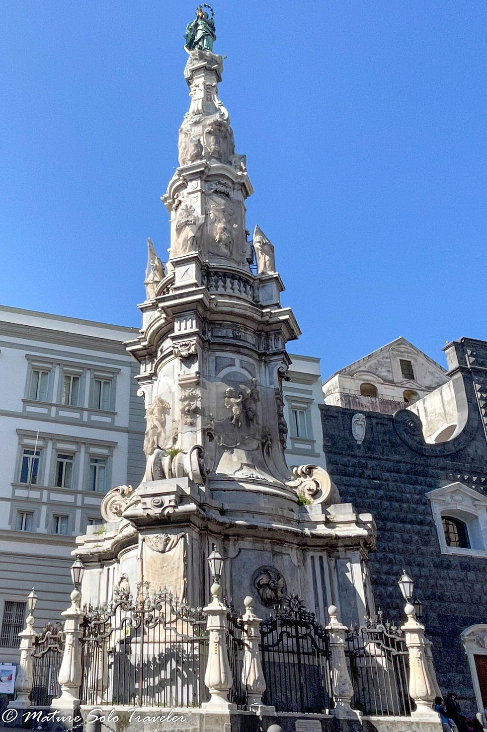 a tall marble plague spire of the Virgin Mary. Her bronze statue stands atop the spire. the four sides are ornately decorated with scenes from her lift. A metal barricade surrounds the spire, to stop anyone touching it.