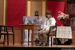 Attore in abiti ottocenteschi è seduto accanto a un tavolino davanti a un camino scenico, in un interno elegante con fiori e arredi d’epoca, in atteggiamento pensieroso.