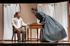 Scena teatrale in costume ottocentesco: una donna in abito scuro si china su un uomo seduto a un tavolino, in un ambiente semplice con paraventi, mentre lo osserva con tono deciso.