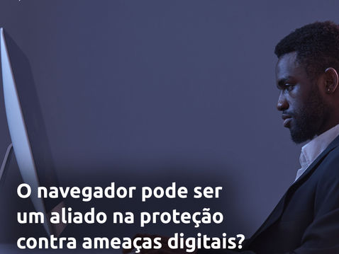 A imagem apresenta um homem negro, de barba e cabelo curto, vestido com um blazer escuro e camisa branca, utilizando um computador de mesa em um ambiente com iluminação escura e tonalidade azulada.