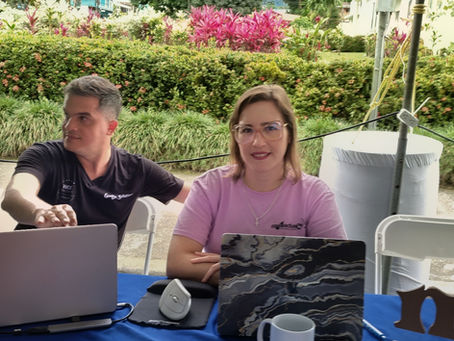 Víctor González Dondi y Melissa Vargas atendiendo un stand de consultoría con laptops sobre una mesa azul, con el majestuoso Volcán Arenal de fondo en La Fortuna, Costa Rica.
