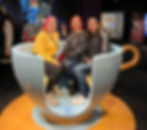 Three people sit in a giant teacup exhibit, smiling. Background features whimsical art and colorful decor. Warm, cheerful mood.