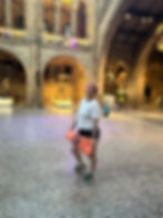 Person in white shirt holds orange yoga mat, smiling in grand hall with arched stone architecture. Skeleton and people in background.