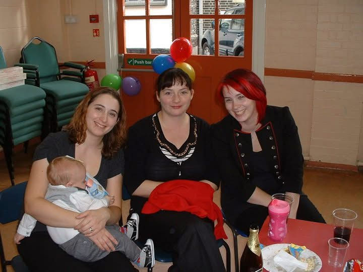 Three women seated indoors; one holds a baby. Balloons decorate a door behind them. A festive, cheerful atmosphere with colorful accents.