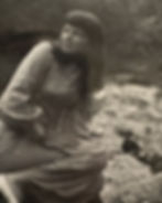 Woman in a checkered dress sits beside a stream in a wooded area, looking upward with a contemplative expression. Black and white photo.