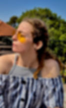 Person with curly hair and yellow glasses sits outdoors, wearing a striped off-shoulder top, looking to the side. Green trees and a house roof in the background.