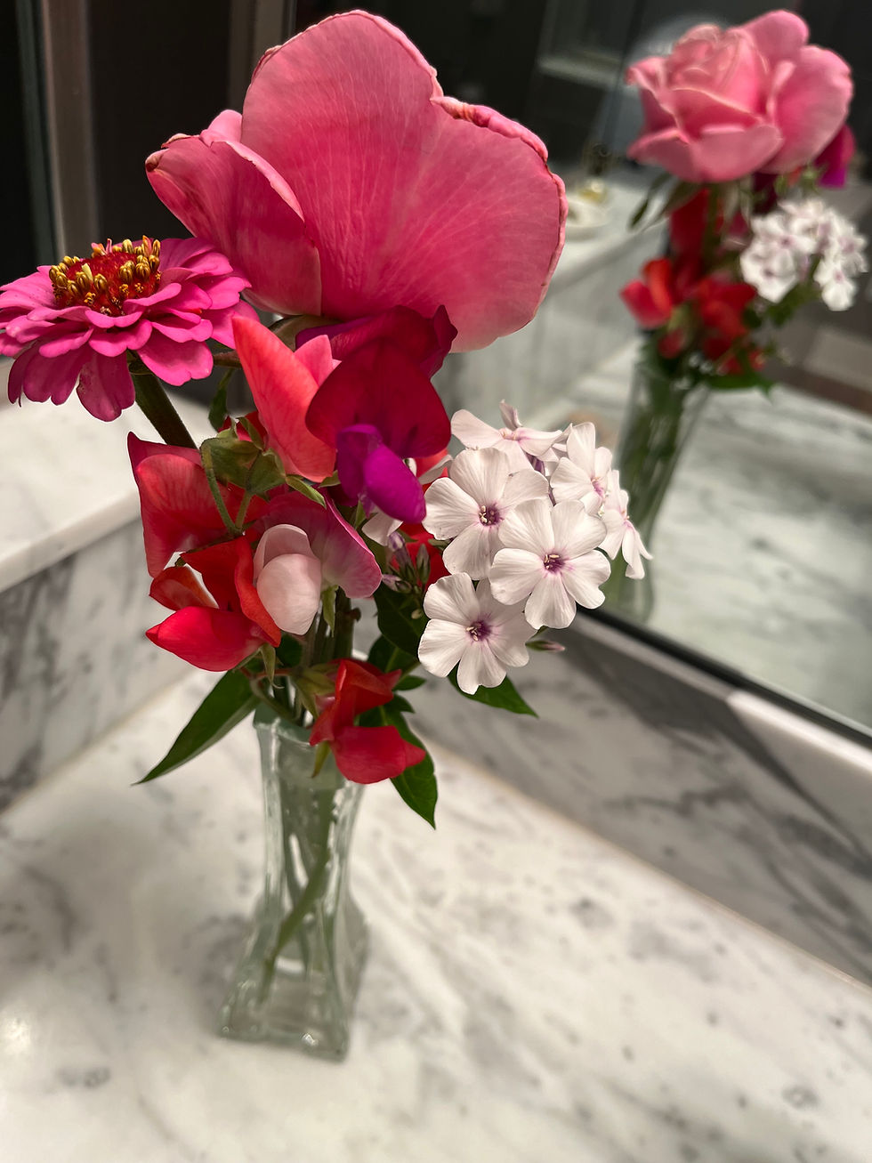 A glass vase with pink and white flowers on a marble countertop, reflecting in a mirror, creating a vibrant and elegant scene.