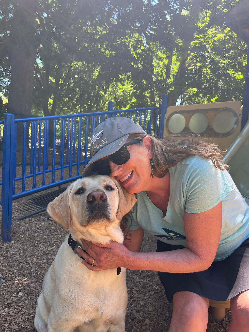 Woman in a blue cap hugs a yellow Labrador in a sunny park. Blue fence and trees in background. Both appear happy and content.