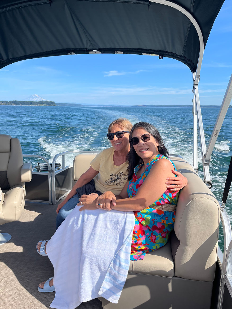 Two women smiling on a boat, seated under a canopy. One wears a colorful dress. Calm water and distant mountains in background, sunny day.