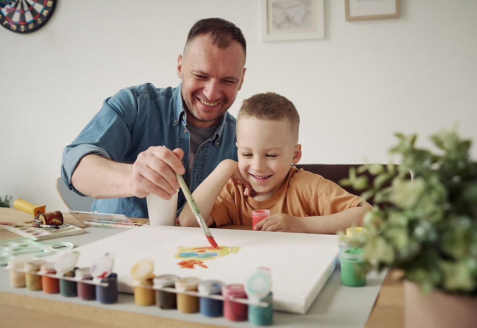 A smiling father paints with his young son at the kitchen table, sharing a quiet, joyful moment that reflects intentional and engaged parenting.