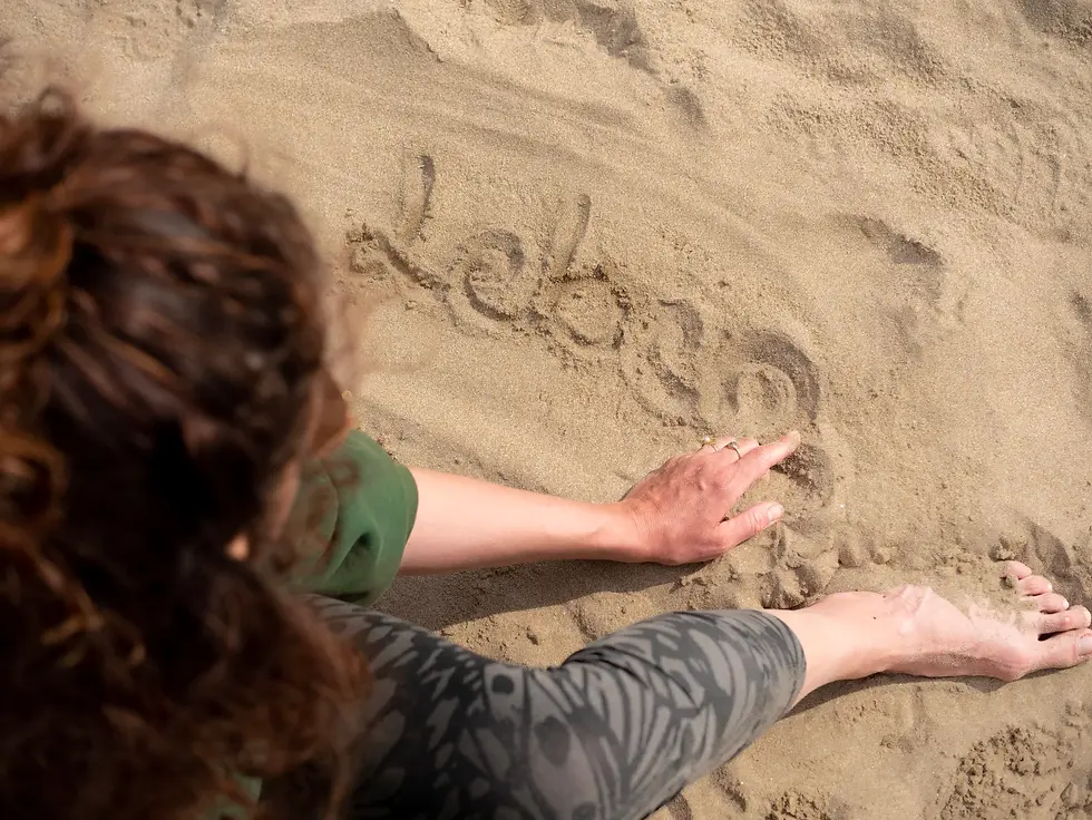 Nahaufnahme aus der Vogelperspektive: Eine Frau mit lockigem Haar schreibt mit dem Finger das Wort ‚leben‘ in den Sand. Man sieht ihre Hand mit Ringen und ihre nackten Füße im Sand.
