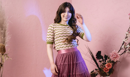 Woman standing wearing a shirt and long pink skirt. The backdrop is all pink.