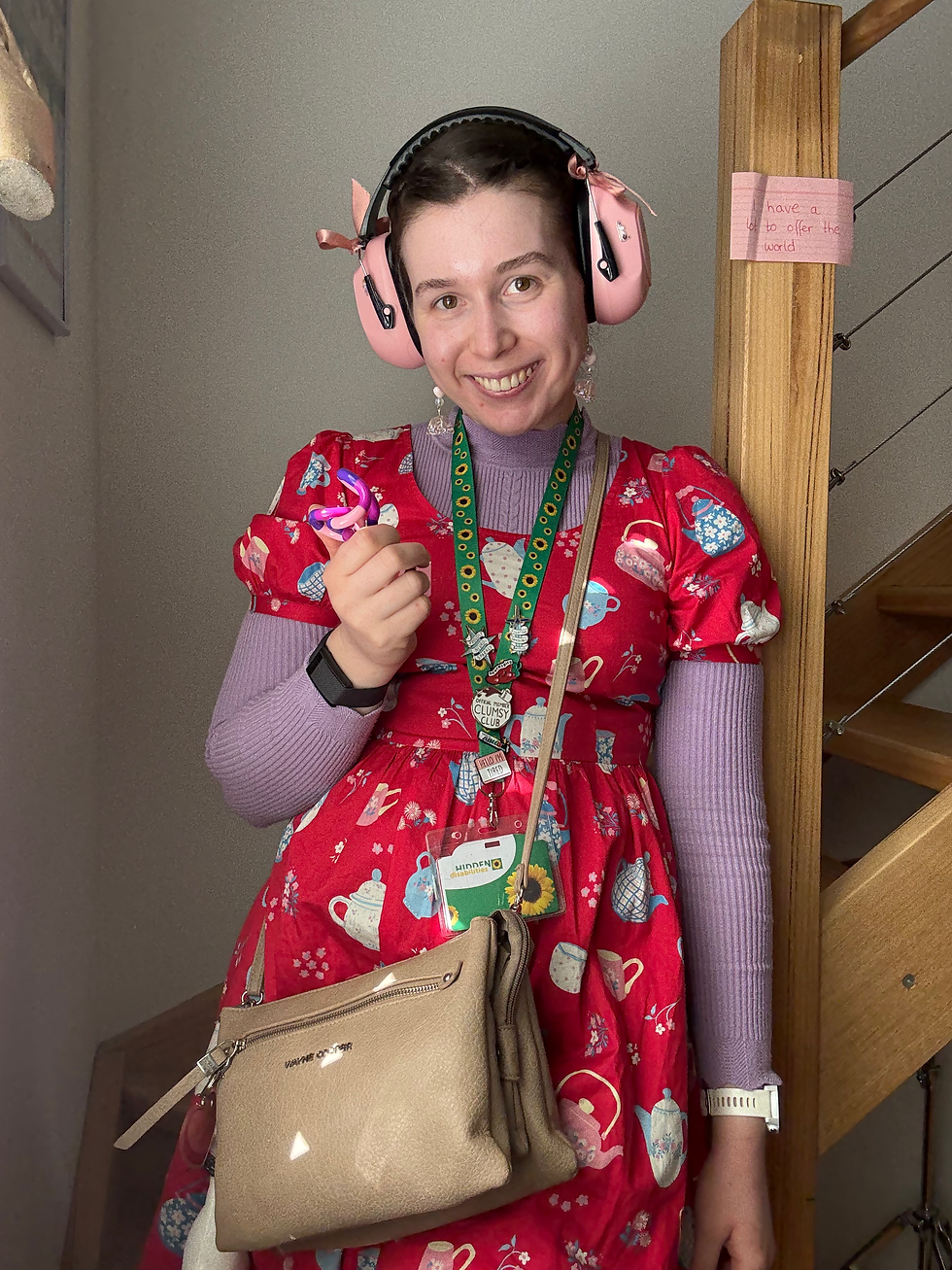Saffron, a young woman, stands against a plain background. she is wearing pink ear defenders and the hidden disabilities sunflower lanyard over a bright red dress. She is holding a tangle fidget toy in her right hand and smiling at the camera.