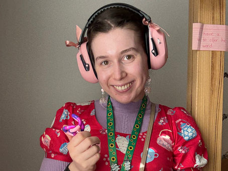 Saffron, a young woman, stands against a plain background. she is wearing pink ear defenders and the hidden disabilities sunflower lanyard over a bright red dress. She is holding a tangle fidget toy in her right hand and smiling at the camera.
