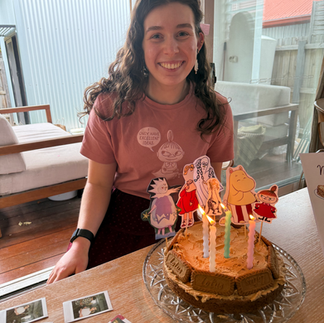 Image of Saffron, a young woman wearing a pink shirt, smiling and sitting behind a birthday cake with the candles lit.