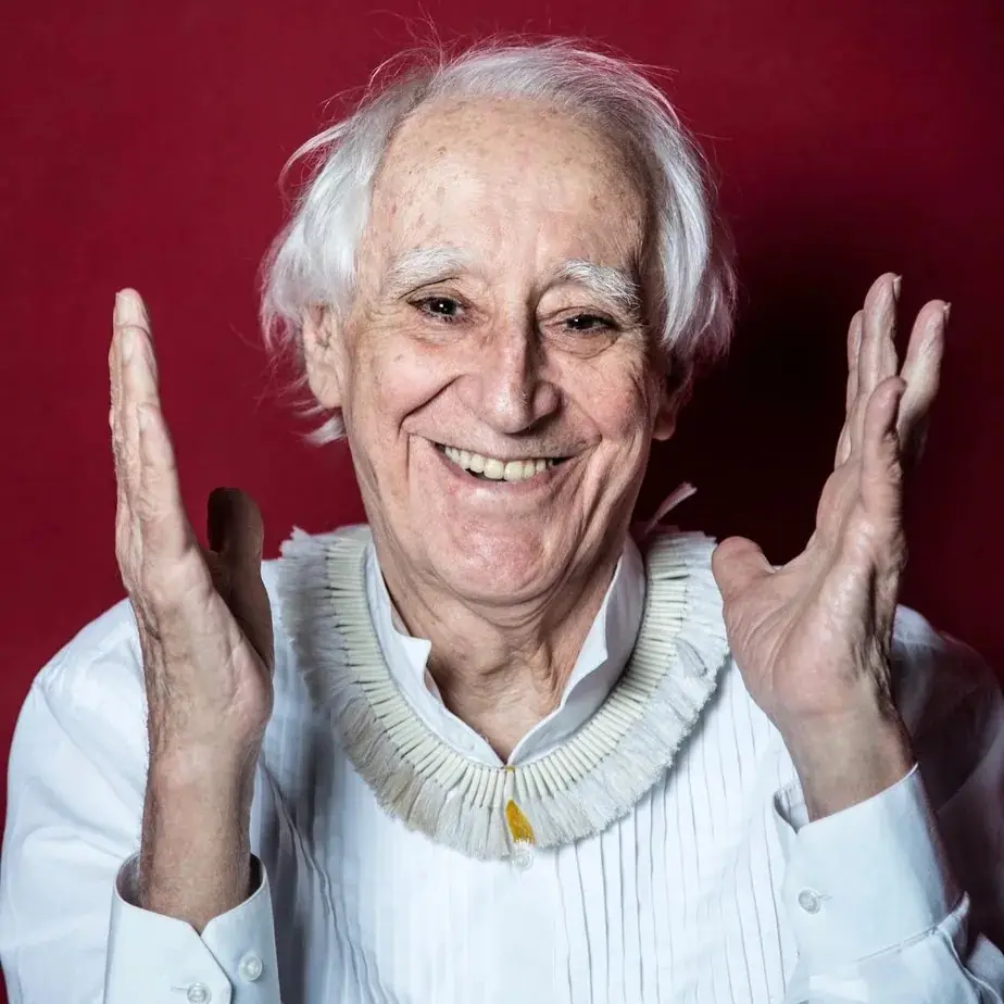 Zé Celso sorrindo, de camisa branca e colar, com as mãos abertas diante de um fundo vermelho