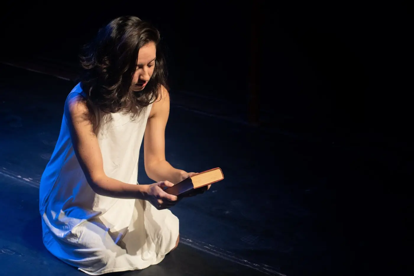 Atriz ajoelhada no palco, lendo um livro sob luz azul