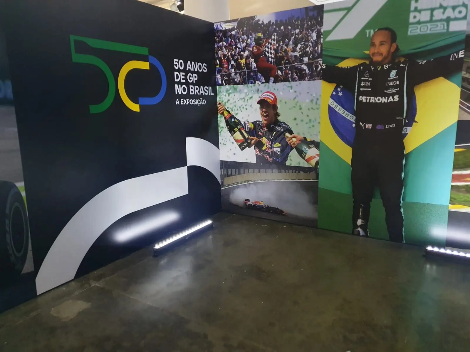 Painel de abertura da exposição com o selo 50 Anos de GP no Brasil, fotos de Sebastian Vettel e Lewis Hamilton com a bandeira do Brasil