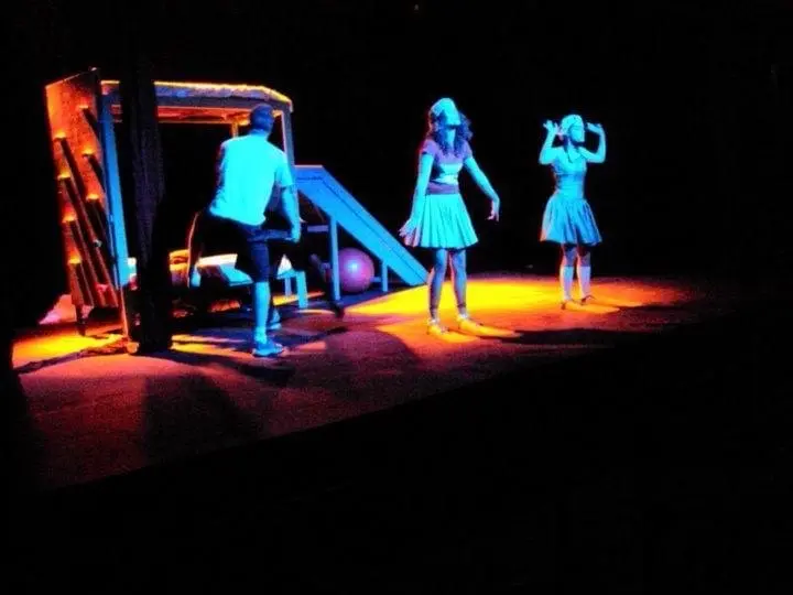 Elenco no palco sob luz azul e laranja, ao lado de um cenário com escorregador