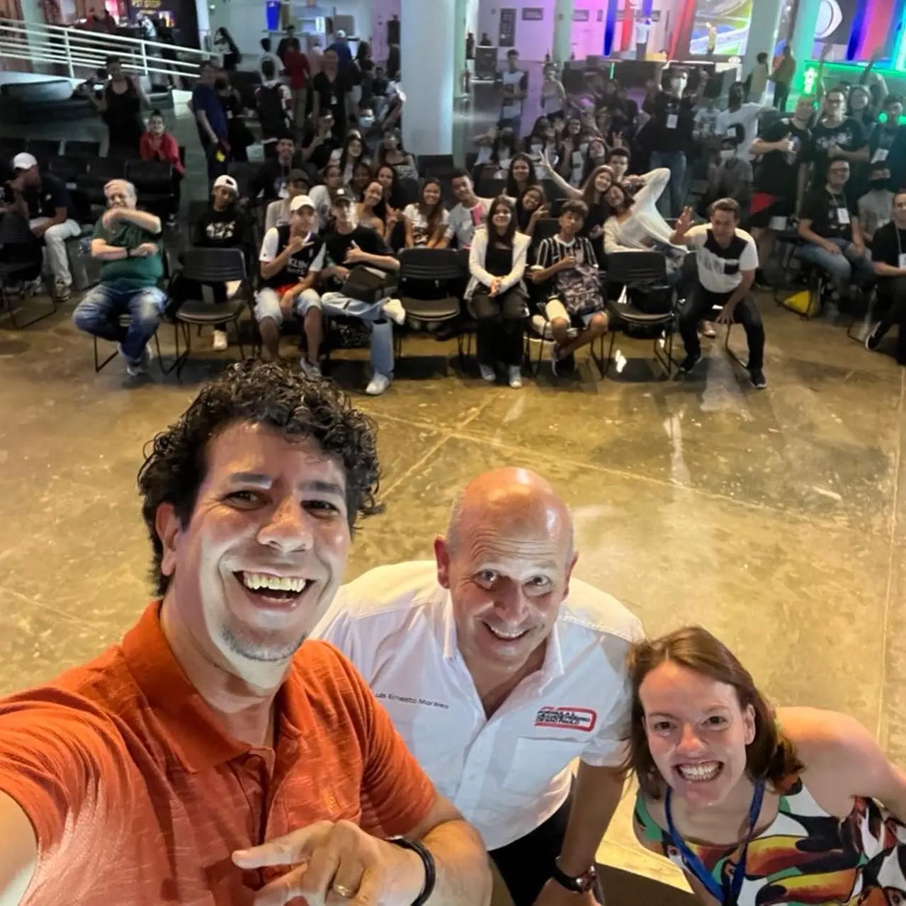 Selfie dos apresentadores e convidado com a plateia de estudantes ao fundo