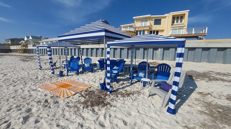 Blue striped canopies and chairs on a sunny beach, with a yellow house in the background. Orange striped mat on sand, relaxed vibe.