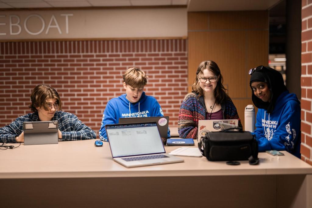 A brown haired boy with glasses sits in front of a iPad looking over at the blond next to him who is looking at his laptop with a surprised expression. Next to the two is a grey haired girl sitting in front of Surface laptop smiling at the camera. The final person at the table is another girl smiling at the camera.