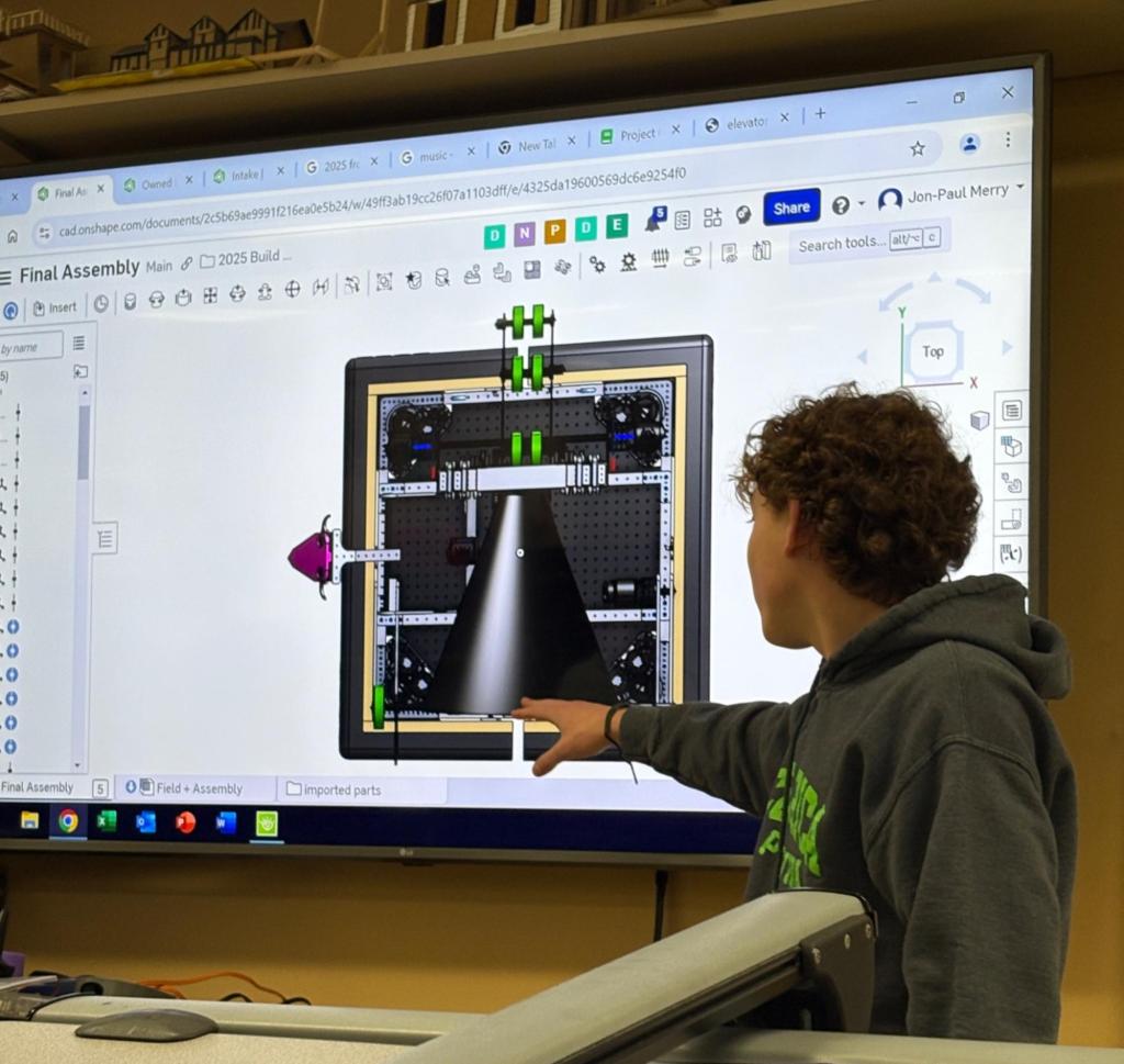 Drew M, wearing a gray hoodie with a lime green design on it, is pointing at a large screen displaying a CAD (Computer-Aided Design) model within the Onshape software interface. The CAD model showcases a detailed assembly of a robot, with various components and parts visible. He is emphasizing a specific section of the model, giving a presentation.