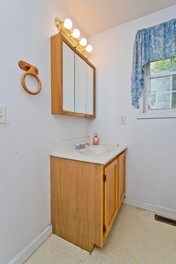 A wooden vanity, white top, and wooden medicine cabinet that has a mirror on the front above the vanity. There is a window and white walls.
