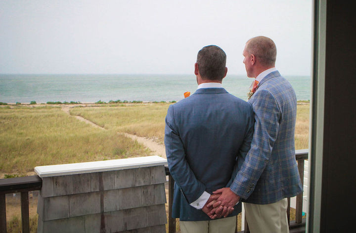 Two grooms dressed in grey blue jackets. Their backs are to you, holding hands. They look over a vast view of sand dunes and ocean.