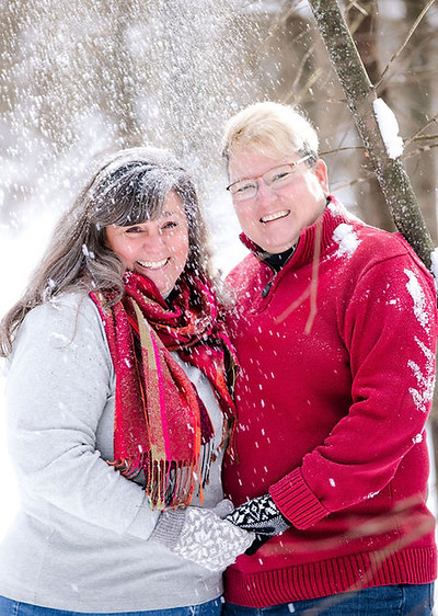 Piper Jo Nevins and her wife Pat hold hands as they look forward, smiling, during a snowing winter photo session.