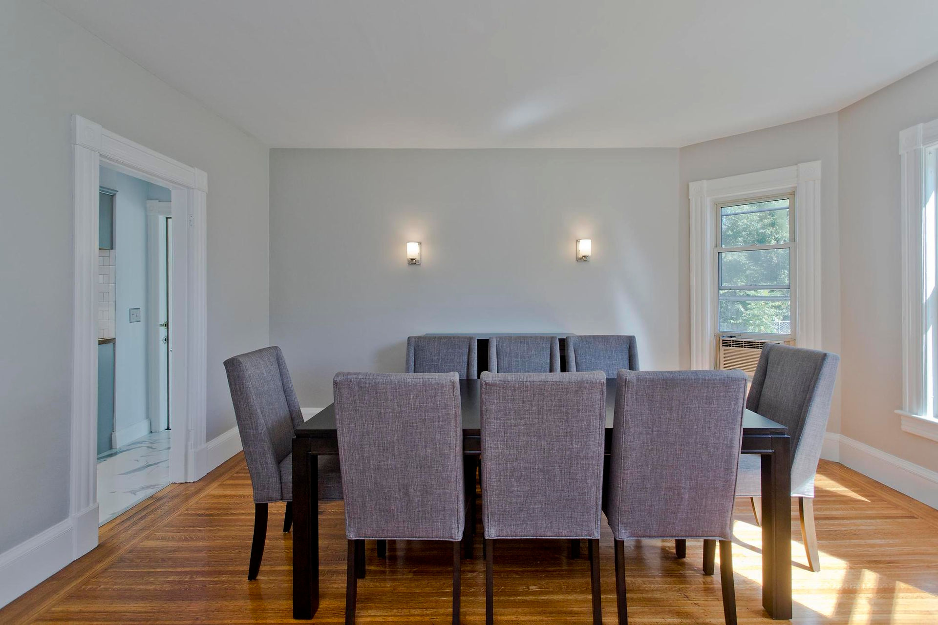 The dining room has wood floors and a large bay window. It opens from a big arch into the living room. There's two sconces on the wall. 